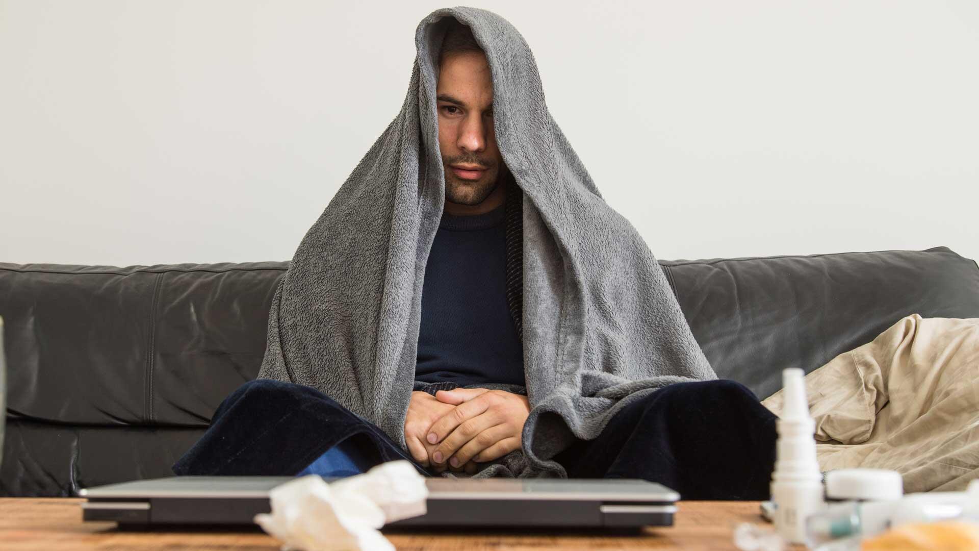 Person unter Decke sitzt auf der Couch, davor Taschentücher.